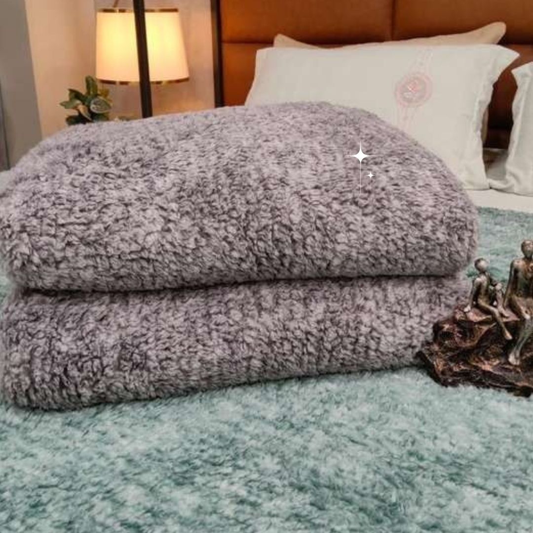 Gray textured blanket on a bed with decorative pillows and a lamp in the background.