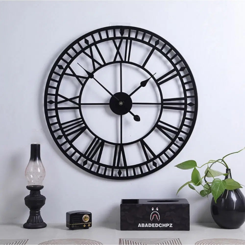 Large black wall clock with Roman numerals on a white wall, accompanied by decorative items.