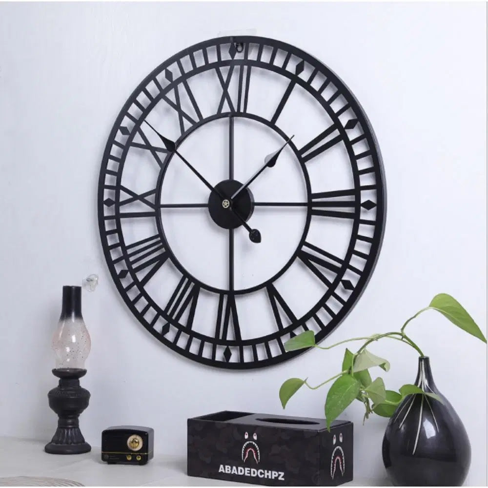Large decorative wall clock with Roman numerals on a white wall, accompanied by a small plant and decorative items.