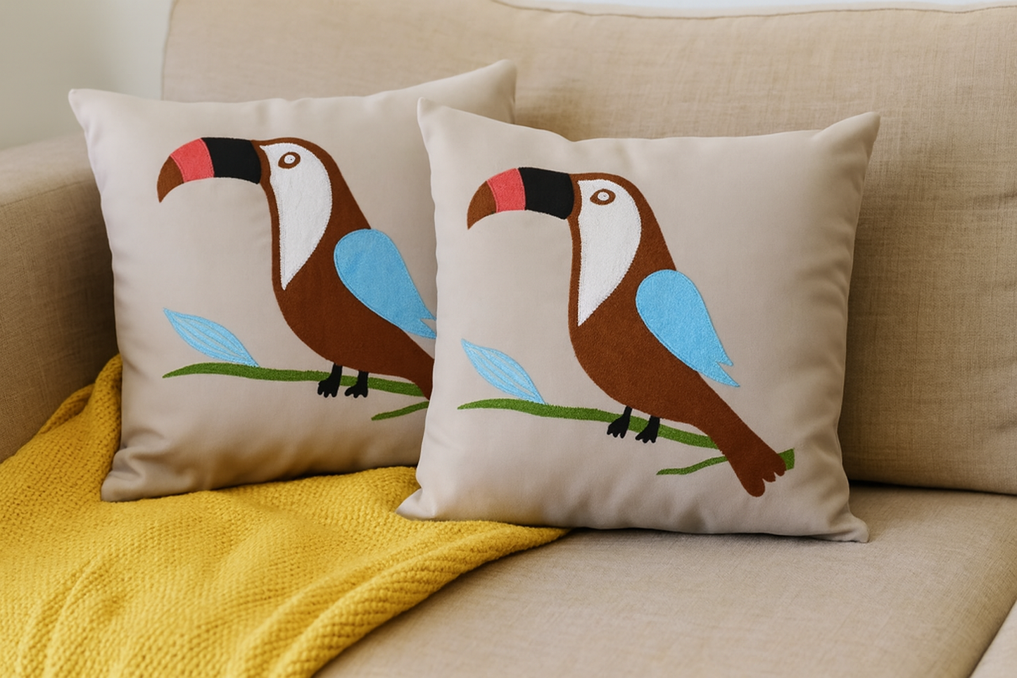 Two decorative pillows with toucan designs on a beige couch with a yellow blanket.