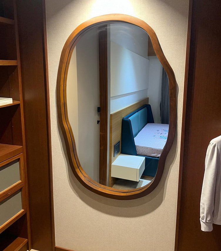 Oval mirror with wooden frame on a wall, reflecting a room with a bed and desk.