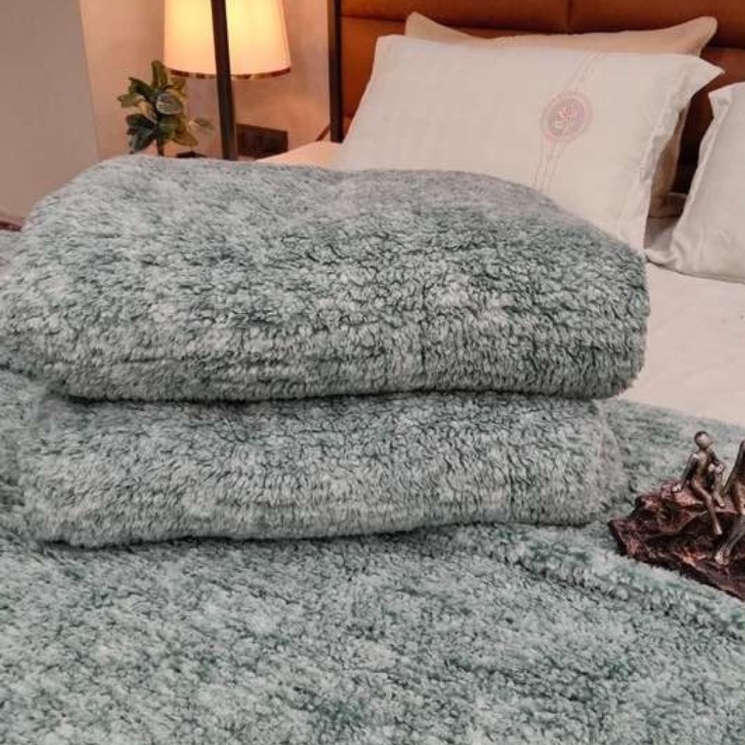 Stack of fluffy green blankets on a bed with pillows and a lamp in the background.