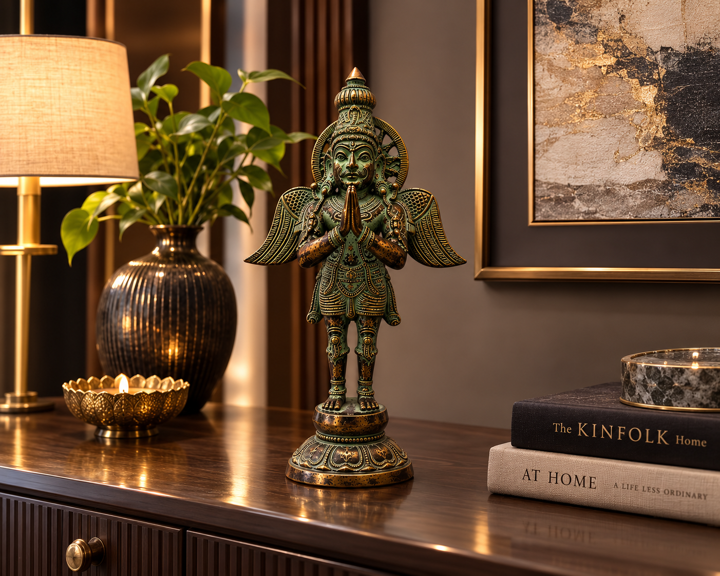 A detailed bronze statue of a winged deity with hands in prayer pose, displayed on a dark wooden console table in a styled room setting with a lamp, plant, and books.
