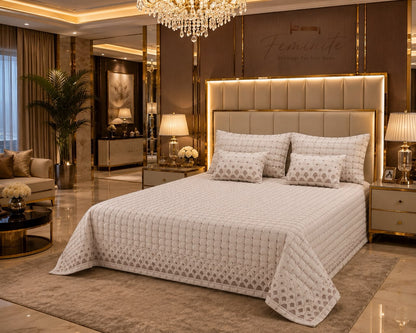 A luxury bedroom scene featuring a bed dressed in a textured cream quilted bedspread and matching pillow shams, set against a modern beige tufted headboard with warm backlighting. The room is styled with gold accents, a crystal chandelier, and a plush rug.
