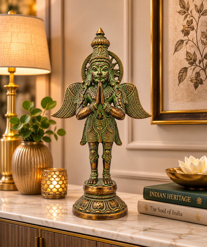 An ornate green and gold Hindu deity statue of a winged figure with a pranamask face, displayed on a marble console table in a styled home decor setting with a lamp, books, and framed art.