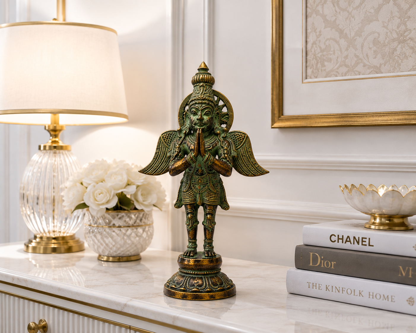 A decorative bronze-toned winged statue of a praying figure stands on a white marble console table in an elegant room setting, styled with a table lamp, white flowers, framed wall art, and a stack of books including Chanel, Dior, and The Kinfolk Home.