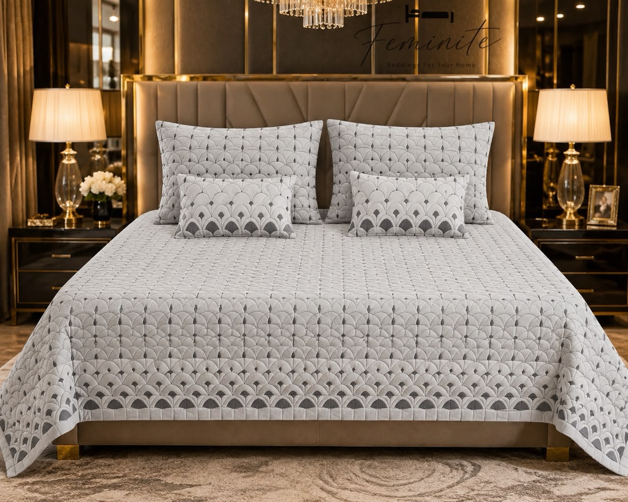 A neatly made bed in a luxurious bedroom setting, featuring a light gray and white geometric patterned quilt, matching pillow shams, and two small accent pillows. The background includes a tufted headboard, bedside tables with lit lamps, and warm ambient lighting.