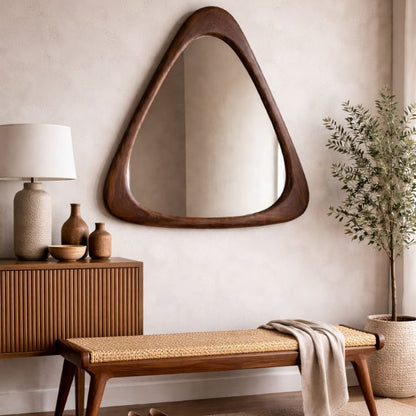 Triangular mirror on a wall above a wooden bench with a lamp and decorative items.