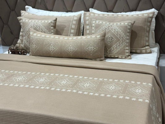 Beige patterned bedspread and pillows on a bed with a brown headboard.