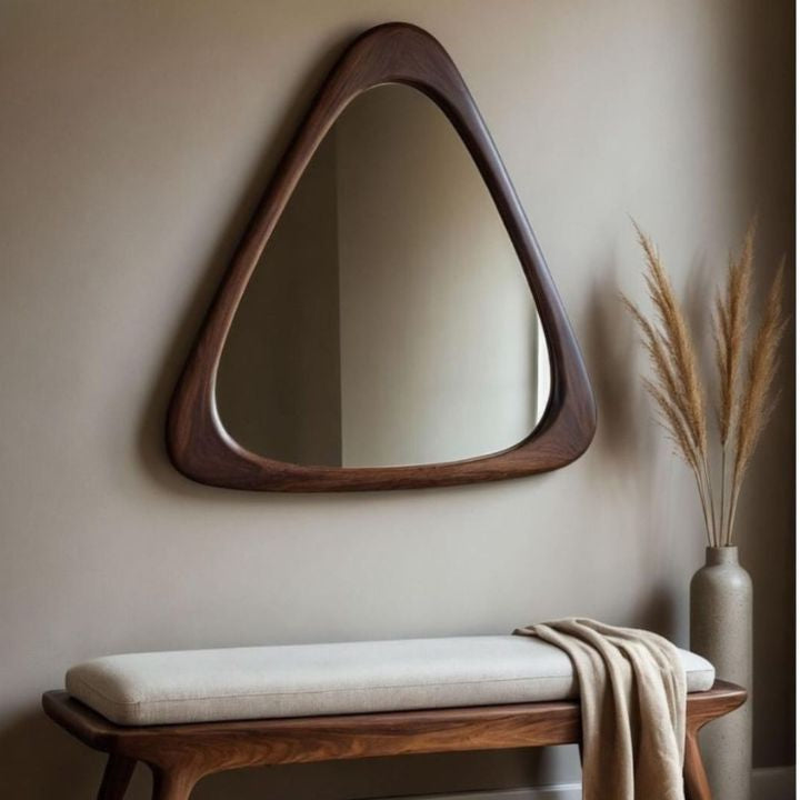 Triangular wooden mirror on a beige wall with a bench and decorative vase below.